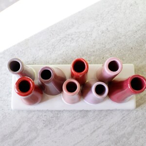 May include: A set of eight ceramic bud vases in a gradient of pink and red hues. The vases are arranged in a row on a white ceramic tray.