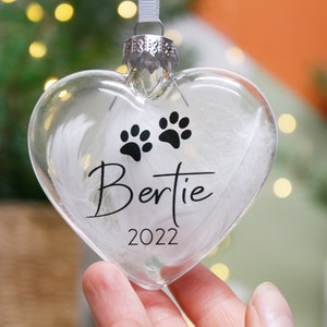 May include: Clear glass heart-shaped bauble with white feathers inside. The bauble has black paw prints and the text "Bertie 2022" printed on it.