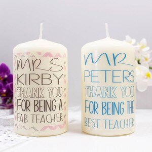Op de afbeelding: Twee witte kaarsen in de vorm van een pilaar met gepersonaliseerde berichten. De kaars links zegt "Mevrouw Kirby Bedankt dat je een fantastische leraar bent" in zwarte en roze letters. De kaars rechts zegt "Meneer Peters Bedankt dat je de beste leraar bent" in zwarte en blauwe letters.