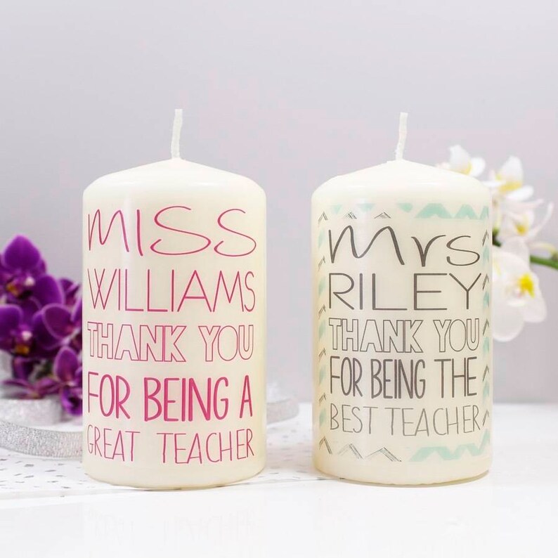Op de afbeelding: Twee witte pilaarvormige kaarsen met gepersonaliseerde berichten. De kaars links zegt "Miss Williams Thank You For Being A Great Teacher" in roze letters. De kaars rechts zegt "Mrs Riley Thank You For Being The Best Teacher" in zwarte letters.