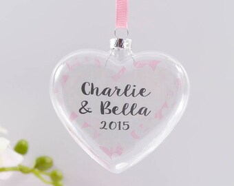 Op de afbeelding: Een transparante glazen hartvormige kerstbal met een roze lint. De kerstbal is versierd met roze glitter en heeft de tekst "Charlie & Bella 2015" erop gedrukt.