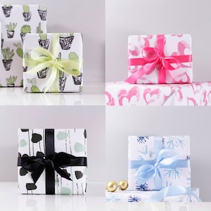 May include: Four gift boxes wrapped in different patterned paper. The boxes are arranged in a grid pattern on a white surface. The top left box is wrapped in white paper with a black and green cactus pattern. The top right box is wrapped in white paper with a pink heart pattern. The bottom left box is wrapped in white paper with a black and green abstract pattern. The bottom right box is wrapped in white paper with a blue snowflake pattern.