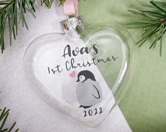 May include: Clear glass heart-shaped Christmas ornament with a penguin and the text "Ava's 1st Christmas 2022".