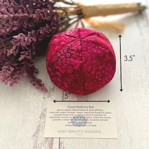 May include: A round, magenta-colored Good Medicine Ball, 5 inches in diameter and 3.5 inches tall. The ball is filled with organic flaxseed, chamomile, and lavender flowers. The product description is printed on a card.