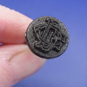 May include: A close-up of a dark gray, round button with a raised, ornate design. The button features a stylized monogram or emblem, possibly for a fashion item. The button is held between fingers against a blue background.