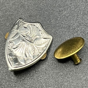 May include: A silver-toned metal shield-shaped button with an engraved floral design and a gold-toned metal button with a round top and a post.