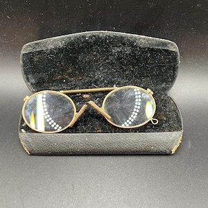 Può includere: Un paio di occhiali rotondi antichi color oro con un motivo decorativo sulle lenti, in una custodia in velluto nero.