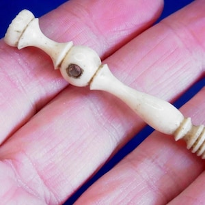 May include: A small, off-white, decorative object with a bulbous center and a threaded end. The object has a cylindrical shape with a series of ridges at one end and a small, flat top. The object is held in a person's hand.