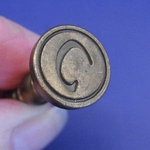 Könnte beinhalten: Ein Vintage-Messingstempel mit kreisförmigem Design. Der Stempel zeigt den Buchstaben "C" in einer stilisierten Schriftart. Der Stempel wird vor einem blauen Hintergrund gehalten. Der Stempel wird wahrscheinlich zum Markieren oder Bedrucken verwendet.