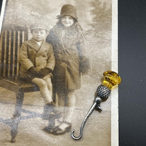 Peut inclure: Photographie en noir et blanc de deux enfants, l'un assis sur un banc, l'autre debout. Une broche argentée avec une pierre jaune ambrée est au premier plan. La broche a un crochet.