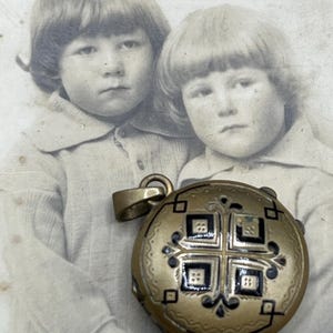 May include: A vintage gold-toned locket with a decorative cross design in black enamel, set against a faded black and white photograph of two children. The locket has a circular shape and a small loop for a chain.