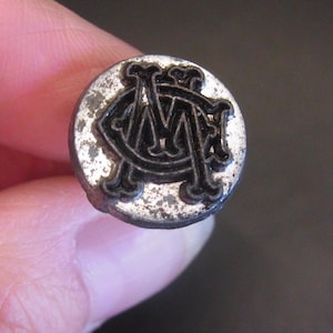 Könnte beinhalten: Ein kleiner, runder, silberfarbener Knopf mit einem schwarzen, verschlungenen "M" und "A" Monogramm. Der Knopf hat ein leicht abgenutztes Aussehen, mit sichtbaren Gebrauchsspuren auf der Oberfläche.