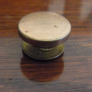 Op de afbeelding: Een gouden metalen knoop met een ronde, licht gewelfde bovenkant en een kleinere, ronde basis.