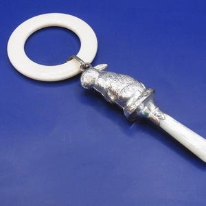 May include: A silver rattle featuring a sculpted rabbit figure attached to a white circular ring and a white handle. The metallic rabbit is textured to resemble fur, and the handle is smooth and cylindrical, connecting to the base of the rabbit.