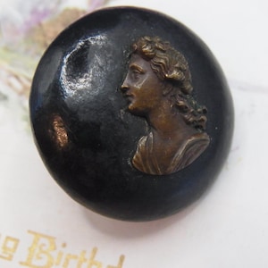 May include: A round, black button with a bronze-colored profile of a person. The button has a glossy finish and a slightly raised relief of the profile. The button is likely vintage and could be used for sewing or as a decorative element.