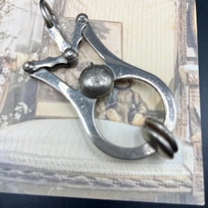 May include: A silver-toned metal chatelaine clip featuring a hinged, scissor-like mechanism with a central circular dome and a small loop at the top. The metal surface is smooth and reflective, with a distinct pivot point and a rounded lower section.