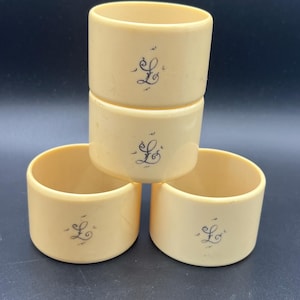 May include: Four cream-coloured napkin rings stacked. Each ring has a black cursive letter "L". The rings are cylindrical with a smooth, matte finish. The background is a dark, solid colour.