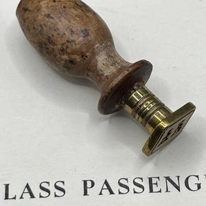 May include: A wax seal stamp featuring a rounded, speckled brown wooden handle attached to a polished brass neck and a square-shaped brass base. The base displays an engraved design. The stamp is positioned above the text LASS PASSENG.