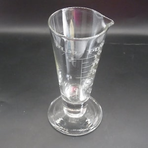 May include: A clear glass measuring beaker with a flared conical shape and a small spout. The beaker has etched measurement markings in fluid ounces and milliliters. It sits on a circular base.
