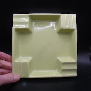 Op de afbeelding: Een vierkante asbak van keramiek met vier inkepingen voor sigaretten. De asbak is lichtgeel.