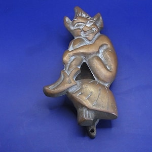 May include: A brass door knocker shaped like a seated figure with pointed ears, wearing a hat and a skirt, with one leg raised. The metal has a warm, brownish-gold tone with visible cast details and a small mounting hole at the bottom base.