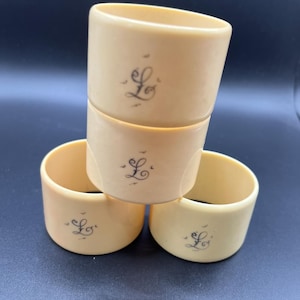 Puede incluir: Cuatro servilleteros de color marfil. Cada anillo presenta una letra "L" en escritura negra con pequeños elementos decorativos. Los anillos están apilados y dispuestos sobre una superficie oscura, mostrando su diseño simple y elegante.
