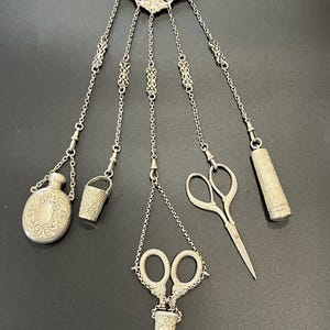 Pode incluir: Uma chatelaine de prata com um clipe superior ornamentado e cinco correntes pendentes. Os acessórios incluem um frasco de perfume oval, um dedal, uma tesoura numa bainha decorativa, uma pequena tesoura de bordar e um estojo cilíndrico.
