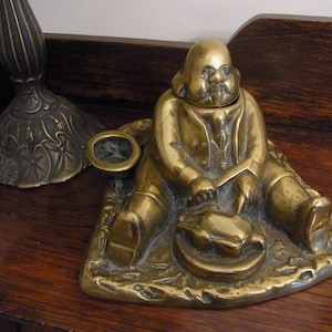 May include: A brass inkwell in the shape of a seated man holding a goose. The inkwell has a round, open top for ink and a small, separate inkwell for a pen.