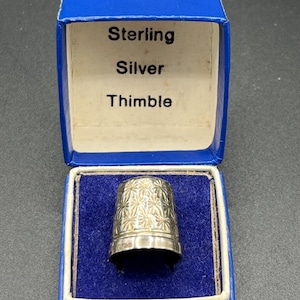 Puede incluir: Un dedal de plata de ley con un diseño floral, presentado en una caja azul. La caja tiene un interior blanco y el texto "Sterling Silver Thimble". El dedal es una herramienta de costura.