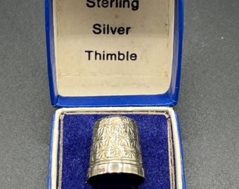 Könnte beinhalten: Ein Sterling Silber Fingerhut mit floralem Muster, präsentiert in einer blauen Box. Die Box hat ein weißes Innenfutter und den Text "Sterling Silver Thimble". Der Fingerhut ist ein Nähwerkzeug.