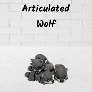 May include: Two grey articulated wolf figurines with visible 3D printed layer lines, black eyes, and black noses. The text "Articulated Wolf" is printed in black above the figures. The wolves have segmented bodies designed for movement.