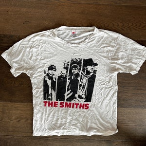 May include: White t-shirt with a black and white image of the band The Smiths. The band members are standing in a row and the band's name is printed in red below the image.