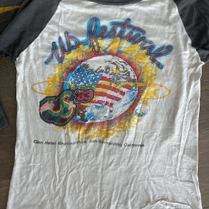May include: A white t-shirt with a faded graphic of a globe with the American flag on it. The text "US Festival" is written in a colorful font above the globe. The text "Glen Helen Regional Park, San Bernardino, California" is written below the globe. The shirt is a baseball style with grey sleeves.