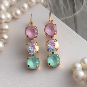 May include: A pair of gold dangle earrings with three crystal stones each. The top stone is pink, the middle stone is iridescent, and the bottom stone is light blue.