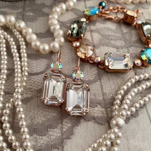 May include: A set of rose gold earrings with clear rectangular crystals and a smaller iridescent crystal on the top. The earrings are displayed on a white pearl necklace.