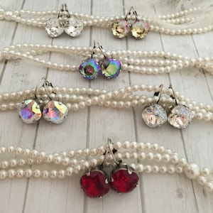 May include: Six pairs of earrings with different colored crystal stones. The earrings are displayed on a white pearl necklace. The stones are iridescent, red, and clear.