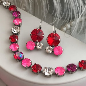 May include: Three necklaces with pink, red, and clear crystal stones. The necklaces are on a white surface with a white feather in the background.