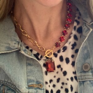 May include: A woman wearing a denim jacket over a white and black animal print top. She is wearing a gold chain necklace with a large red gemstone pendant and a red beaded necklace. She is also wearing red gemstone earrings.