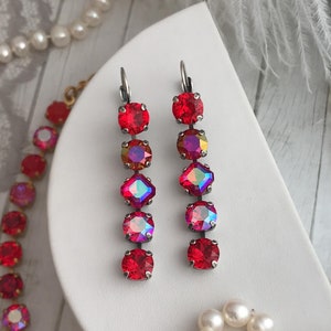 May include: A pair of red crystal earrings with a silver tone finish. The earrings are made up of five crystals each, with the top crystal being the largest and the bottom crystal being the smallest. The crystals are iridescent and have a rainbow effect.