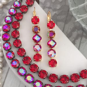 May include: A red rhinestone necklace and matching earrings set. The jewelry features round and square red rhinestones with an iridescent sheen, set in a bronze-colored metal. A pearl necklace is in the background.