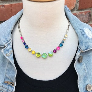May include: A silver chain necklace with rainbow colored crystal stones. The necklace is on a white mannequin wearing a blue denim jacket and a black shirt.