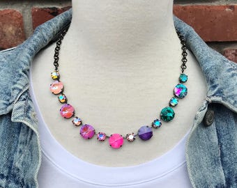Pode incluir: Um colar colorido com pedras preciosas redondas e iridescentes em tons de rosa, roxo, laranja e verde-azulado. O colar é exibido em um manequim vestindo uma camiseta branca e uma jaqueta jeans. A corrente é escura.