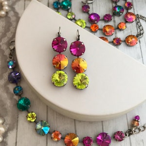 May include: A pair of dangle earrings with three round iridescent crystals in shades of pink, orange, and yellow. The earrings are on a white surface next to a rainbow crystal necklace.
