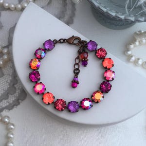 May include: A colorful bracelet featuring a series of round, iridescent gemstones in shades of pink, orange, and purple. The bracelet has a bronze-colored chain and clasp, and is displayed on a white, curved surface.