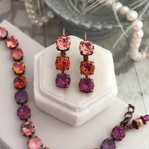 May include: A jewelry set featuring a bracelet and earrings with a gradient of pink, orange, and purple crystals. The bracelet and earrings have a bronze-colored metal setting. The earrings are displayed in a white velvet box.