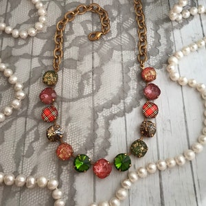 May include: A gold chain necklace with a variety of colorful glass beads. The beads are red, green, and gold with a plaid pattern. The necklace is on a white lace background.