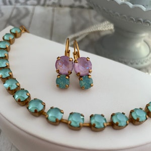 May include: A set of delicate earrings and a necklace featuring light blue and lavender glass stones. The earrings are gold-toned and have a drop design. The necklace is a chain with square-shaped stones.