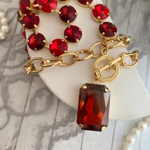 May include: A gold chain necklace with a large red gemstone pendant. The chain is adorned with a double row of smaller red gemstones.