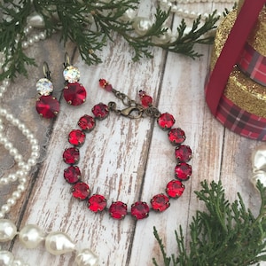May include: A red crystal bracelet with a gold clasp and matching earrings. The bracelet and earrings are on a white wooden background with a pearl necklace and green foliage.