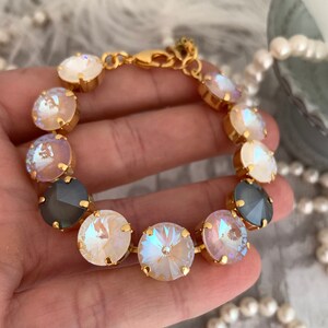 May include: A gold-toned bracelet featuring iridescent, round crystals in shades of white, pink, and gray. The crystals are set in gold bezels and connected by a gold chain. The bracelet is displayed against a soft, blurred background.